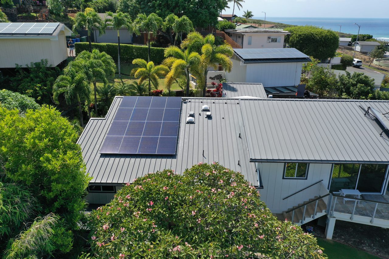 Aerial view of solar panels on a standing-seam metal roof at an Oʻahu home, with tropical greenery and the ocean beyond.